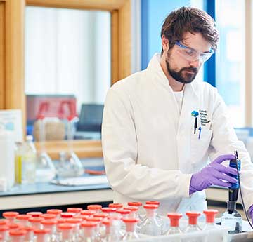 Male student in white personal protective equipment and goggles with purple gloves puts substance into tubes