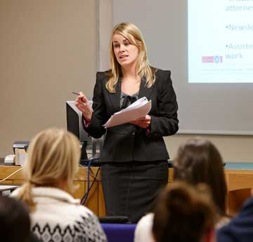 Lady in black suit holding a pamphlet talks to group of students