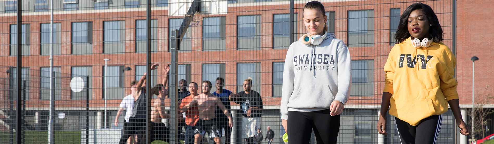 Two students chatting and walking outside a Muga pitch at Bay Campus