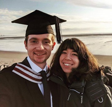 Becky and Joe at Joe's graduation. He is dressed in robes and a mortar board on the beach,