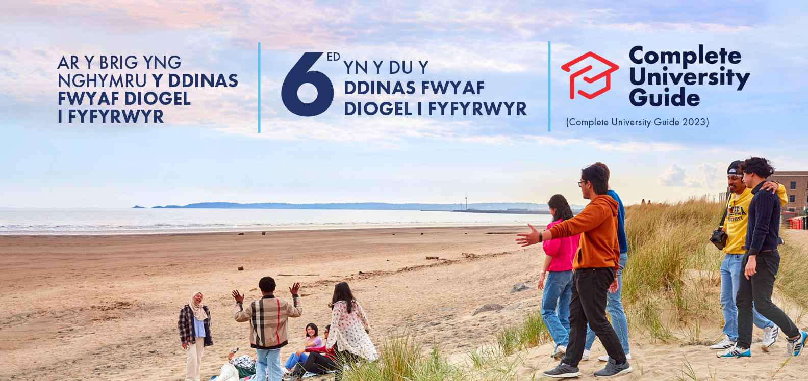 Grŵp o fyfyrwyr yn cerdded ar y traeth a logo Complete University Guide. Testun: Y Ddinas Fwyaf Diogel i Fyfyrwyr yng Nghymru (Complete University Guide 2023) | 6ed yn y DU o ran Diogelwch Myfyrwyr (Complete University Guide 2023)