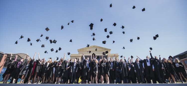 Mae grŵp mawr o fyfyrwyr mewn gwisgoedd graddio'n taflu eu capiau academaidd yn yr awyr wrth sefyll o flaen Neuadd Fawr Prifysgol Abertawe. 