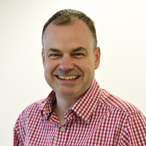 David Smith smiling at camera in a red checked shirt. Neutral background. 