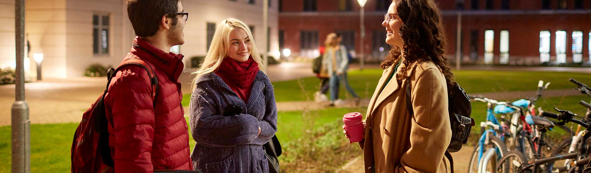 Three students chatting outside Halls of Residence on Bay Campus. The Halls of Residence are lit up by the street lighting.