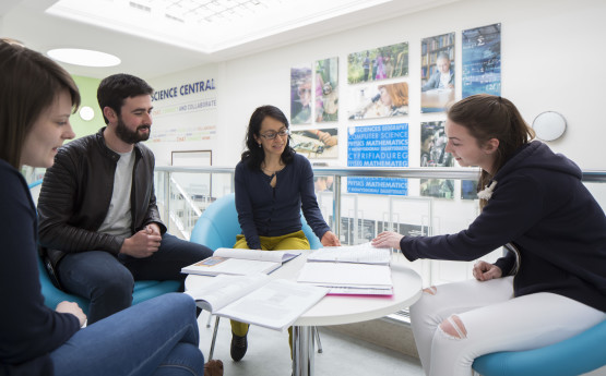 A group of male and female students sat on blue chairs around a white table covered in books, studying together.