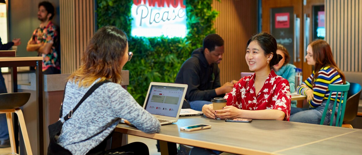 two students on a table talking, with one sitting next to an open laptop