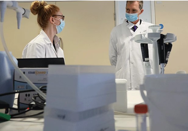 Female scientist in lab wearing blue face mask talking with male in white lab coat and blue face mask