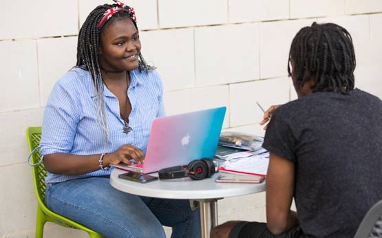 Two students sat outside at a bistro table with a laptop and phones, chatting