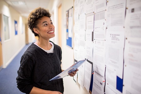 Female student holding a clipboard and looking at a noticeboard on wall.