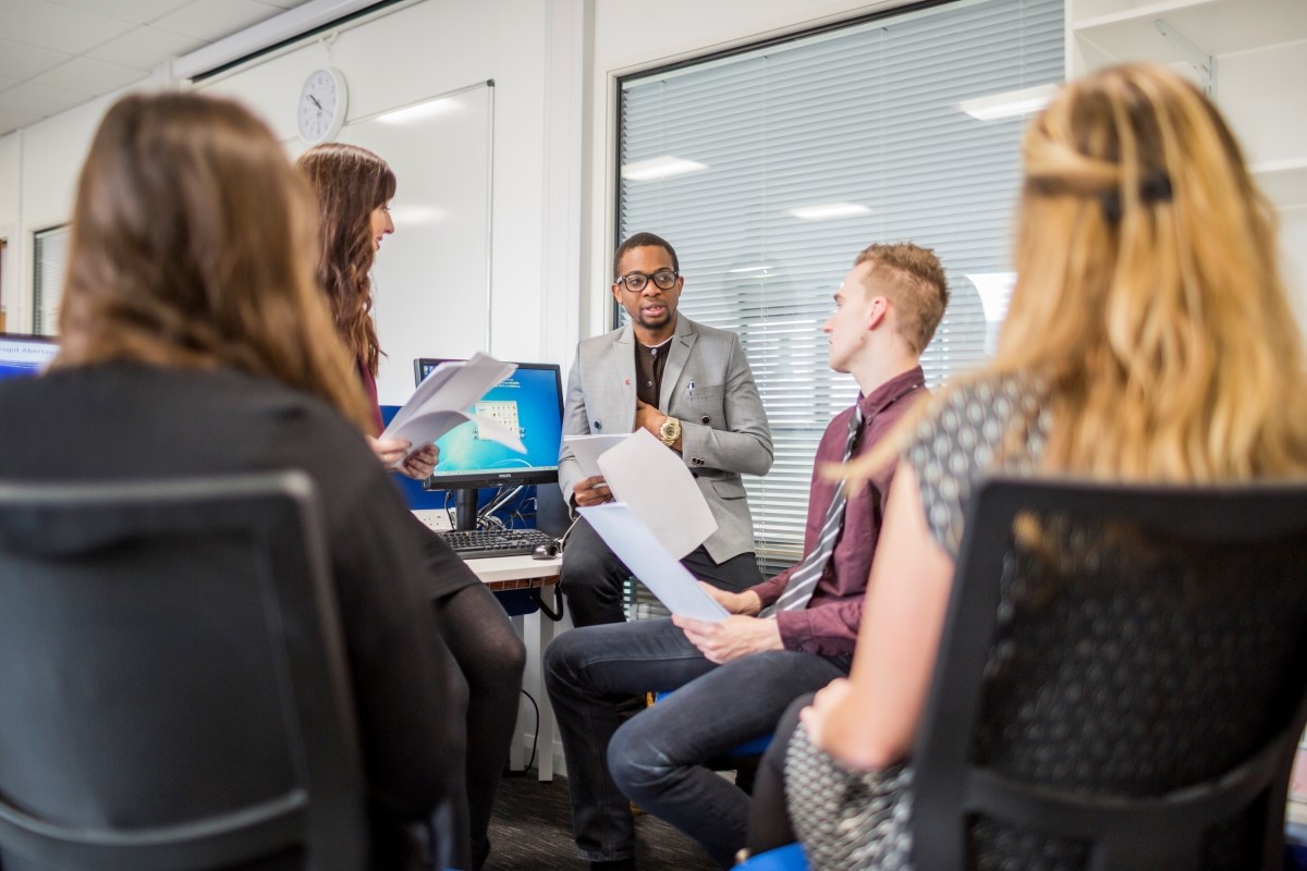 Group of people sitting on chair with one person sitting on table and holding paperwork.