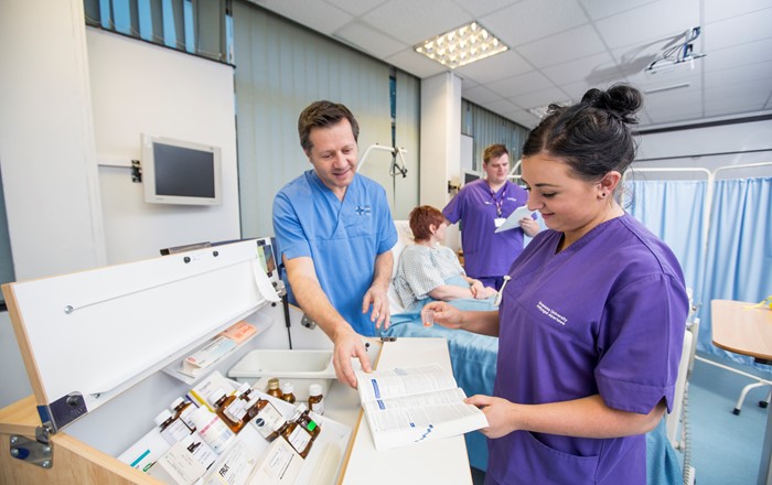 Female student nurse looking at a book under supervision of her teacher while standing by a drug cabinet in a hospital ward. In the background is another male student nurse with a patient in a hospital bed.