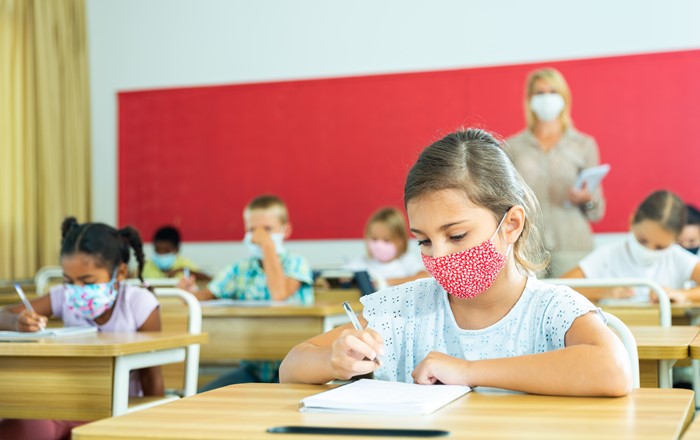 A young schoolgirl wearing a face mask sits writing at her desk, with the teacher and other pupils in the background. 