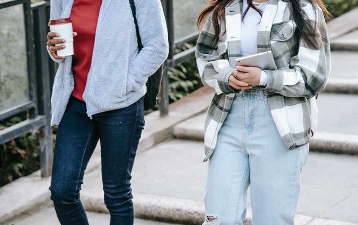 Two teenagers pictured from shoulders down walking side by side down steps, one holding a coffee, one holding a tablet