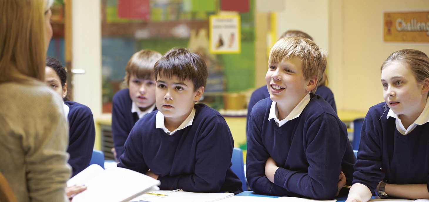 Teacher sitting facing a group of school pupils in uniform who are listening to her reading from a book on her lap. 