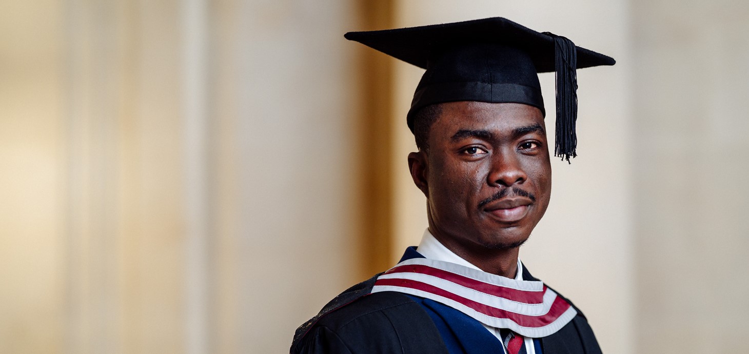 A photo of Martin Tarkpor on his graduation day. Martin is wearing a blue suit and his graduation cap and gown.