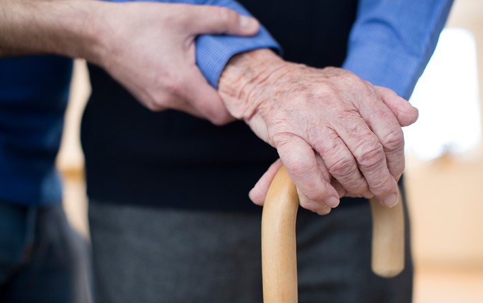 An elderly persons hands on a walking stick with another person's hand offering support