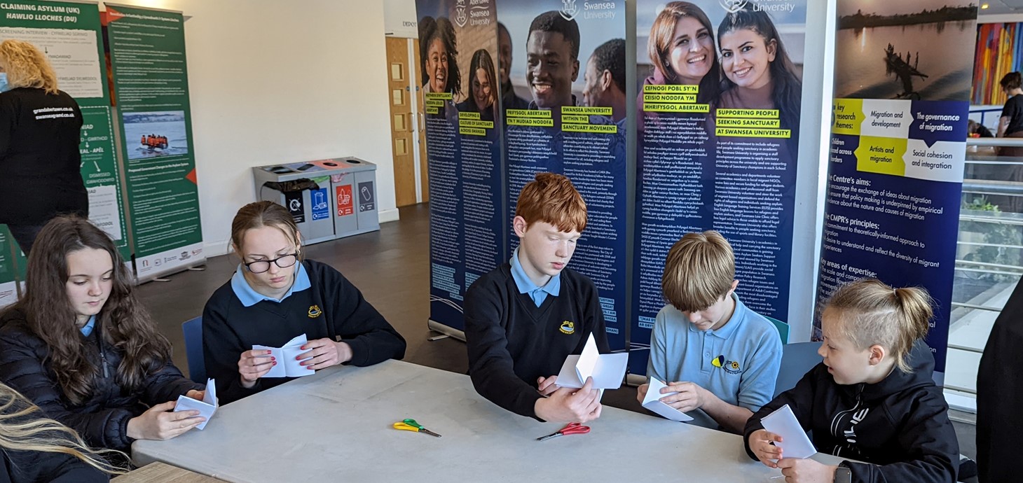 Five school pupils in uniform sitting at a table making things with paper and scissors. Behind them are information panels. Pupils from Ysgol Gyfun Gŵyr in Gowerton taking part in activities at one of the University’s workshops at the exhibition. 