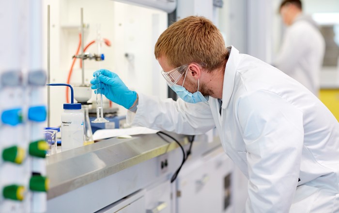 A person wears a surgical mask, white lab coat and gloves as they work in a chemistry lab.