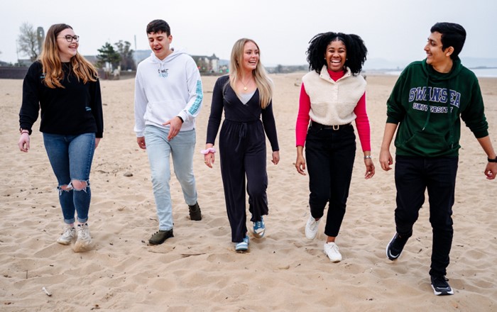 A group of five male and female students are laughing together as they walk on a beach. 