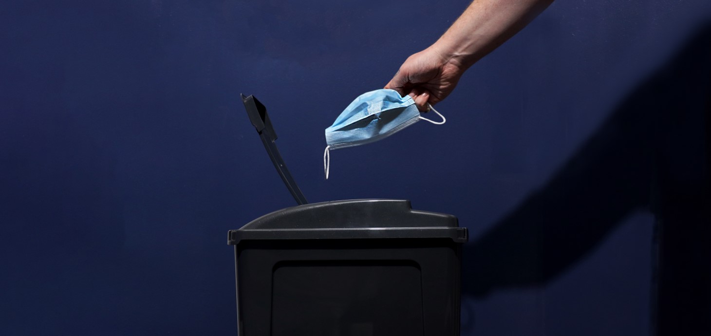 Disposable blue facemask being discarded into black bin.