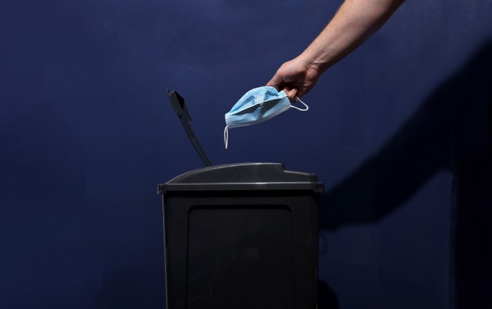 Disposable blue facemask being discarded into black bin.