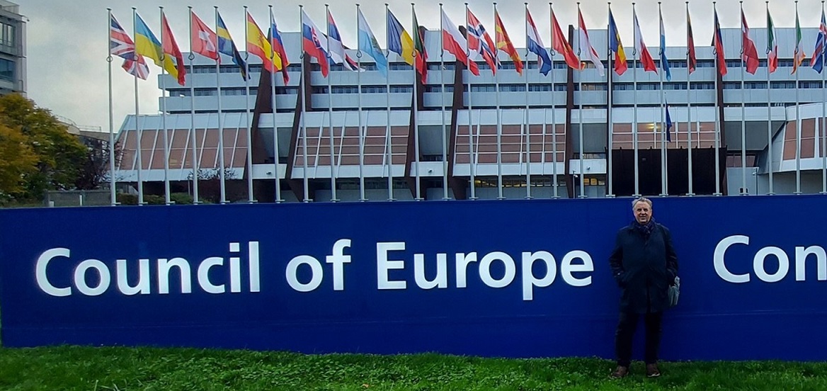 Man standing in front a building with national flags flying behind him.