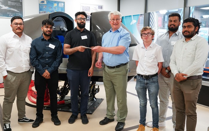 Group of students lined up in front of a flight simulator receiving a cheque from a man.