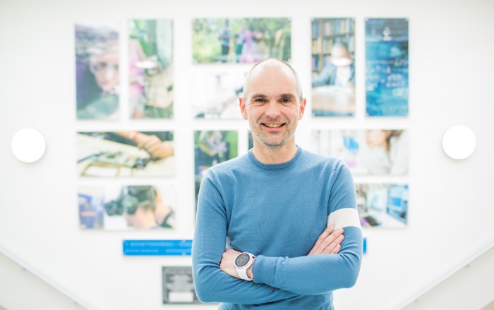 Professor Matt Jones is wearing a blue sweater and is standing in front of a wall of multi-coloured pictures.