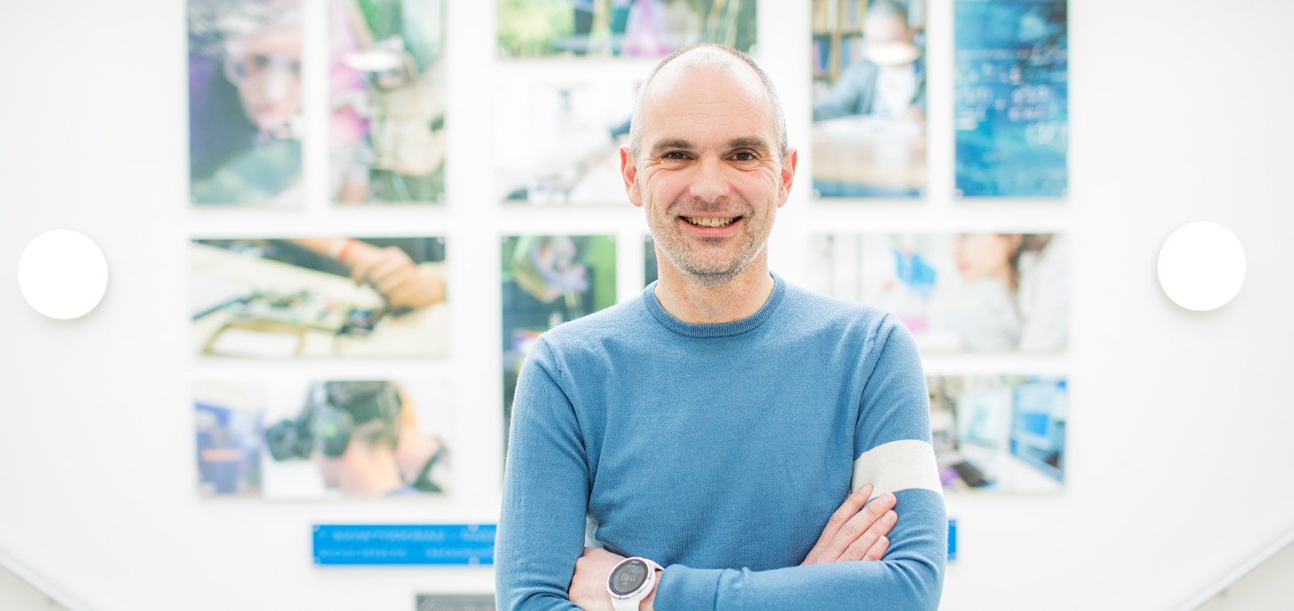 Professor Matt Jones is wearing a blue sweater and is standing in front of a wall of multi-coloured pictures.