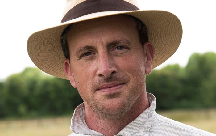 Dr Alexander Langlands is shown in a head and shoulders picture wearing a white shirt with thin blue stripes and a straw hat with a brown band around it.