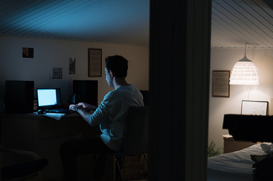 A young man alone in his darkened bedroom sat at a desk looking at his laptop