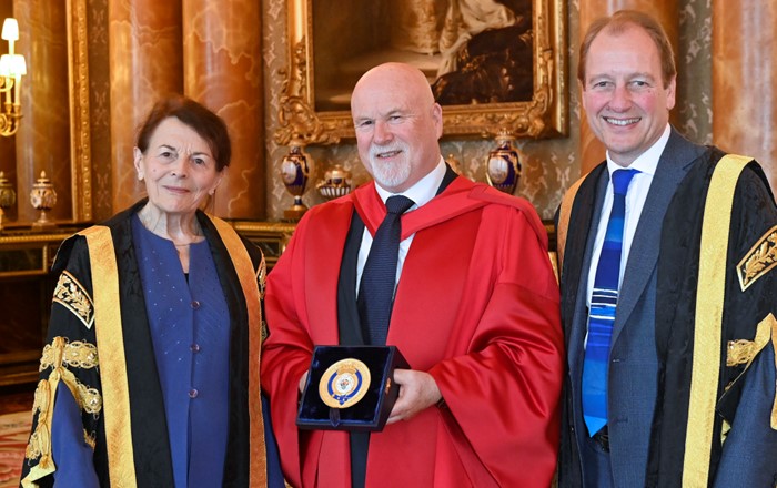 One woman and two men wearing academic robes inside an ornate room. Man in centre of group holding a plaque