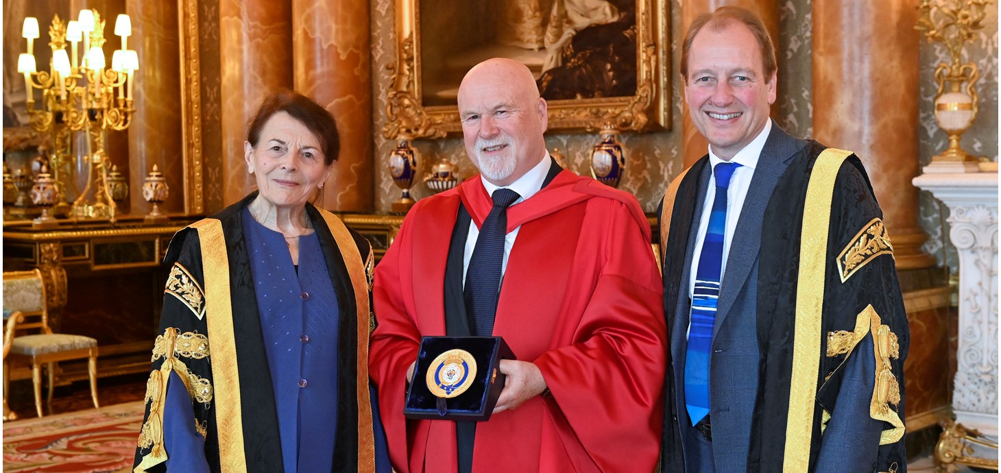 One woman and two men wearing academic robes inside an ornate room. Man in centre of group holding a plaque