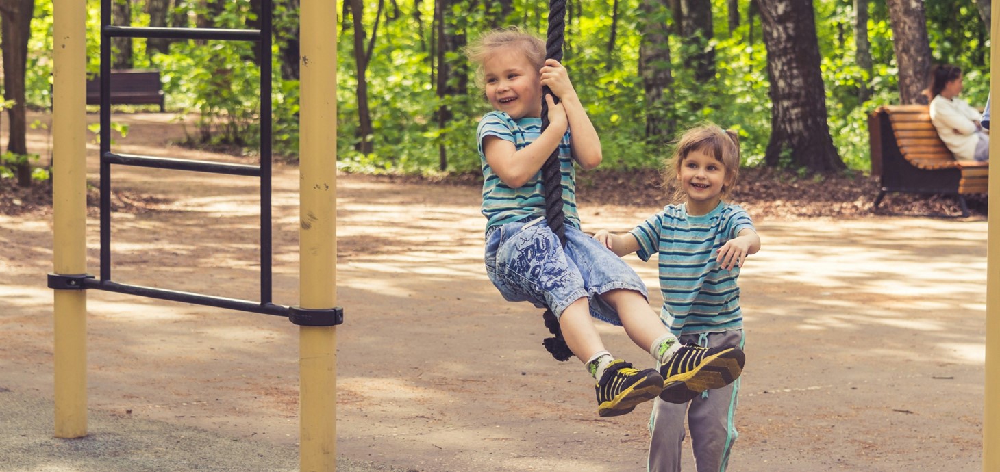 Small girl swinging on a rope in an outdoor playground as another small girl watches