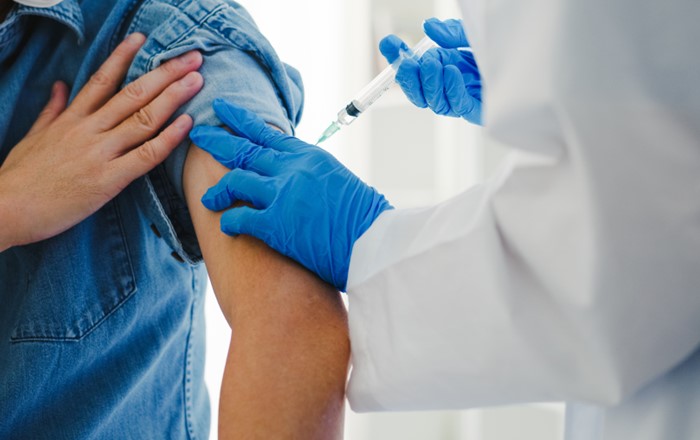 Person in white coat wearing gloves giving an injection into the left arm of another person