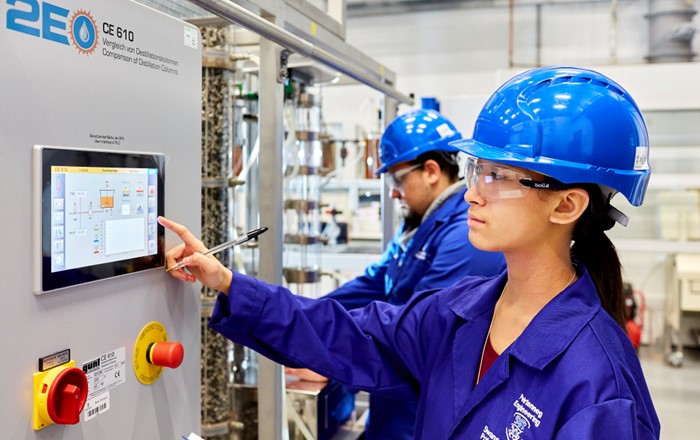 A female student wearing a blue hard hat and overall using a touchscreen computer