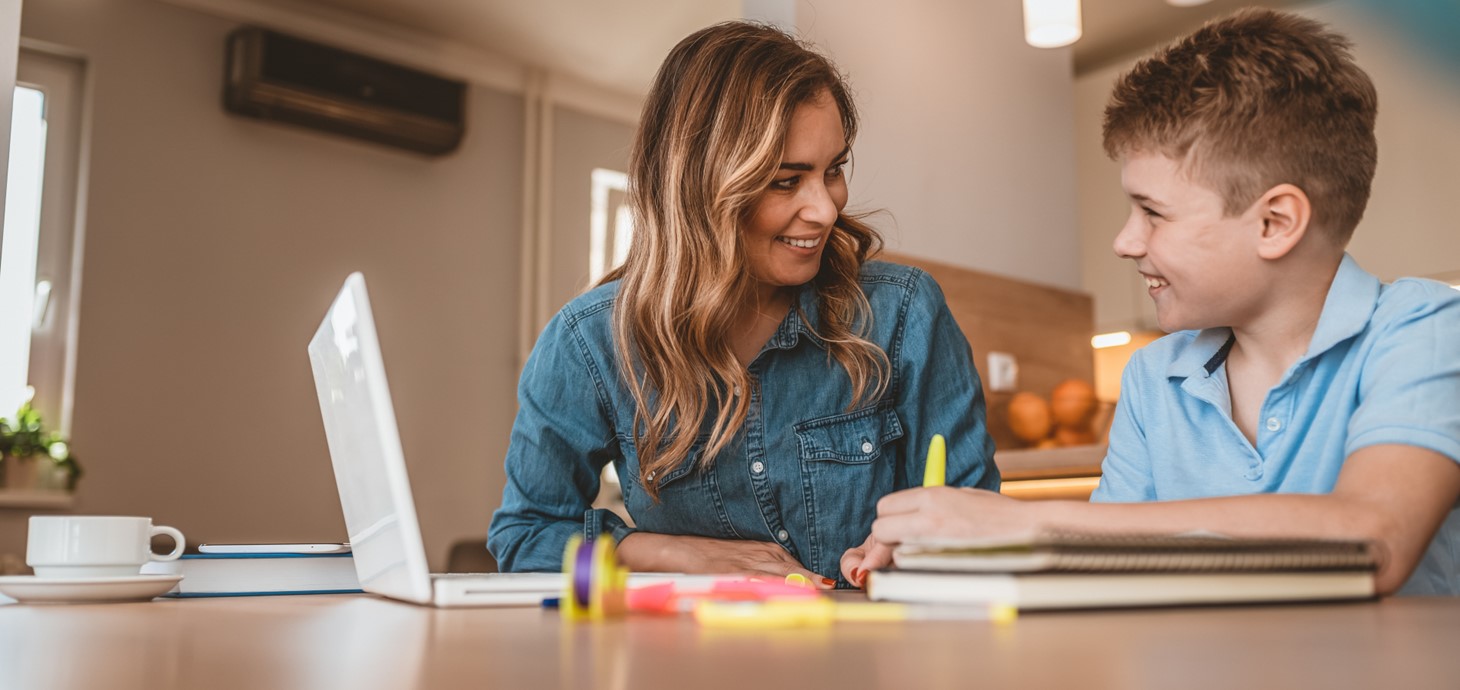 Woman sitting at table smiling at a boy sitting alongside her using a laptop