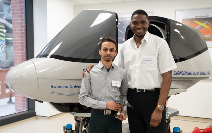 Two young men, one holding a trophy, standing in front of a flight simulator.