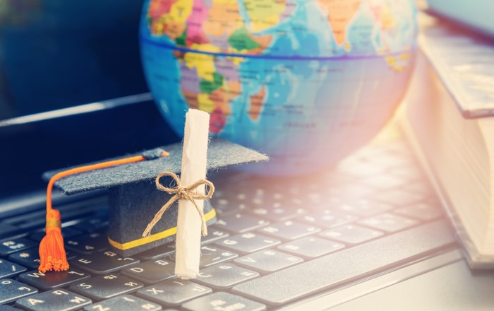 World globe and book in background and a computer keyboard in the foreground with a miniature mortarboard and degree scroll resting on it. 