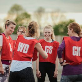 Two students walking outside a Muga pitch at Bay Campus with a game of basketball taking place in the background.