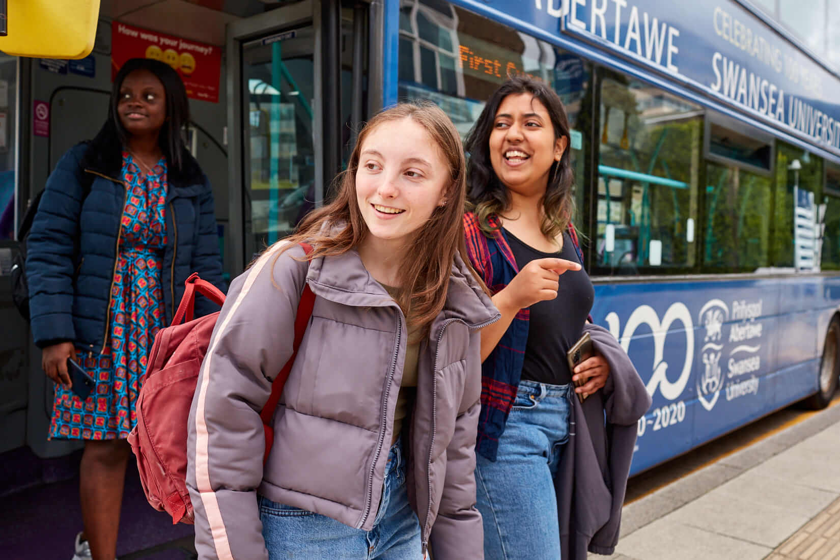 A male and female student looking at a mobile phone. In the background is a First Cymru bus.